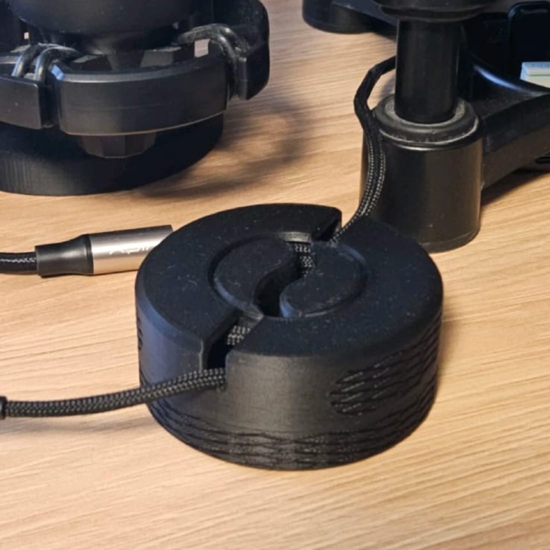 A black 3D-printed circular cable winder on a wooden desk with a braided microphone cable wound into it.