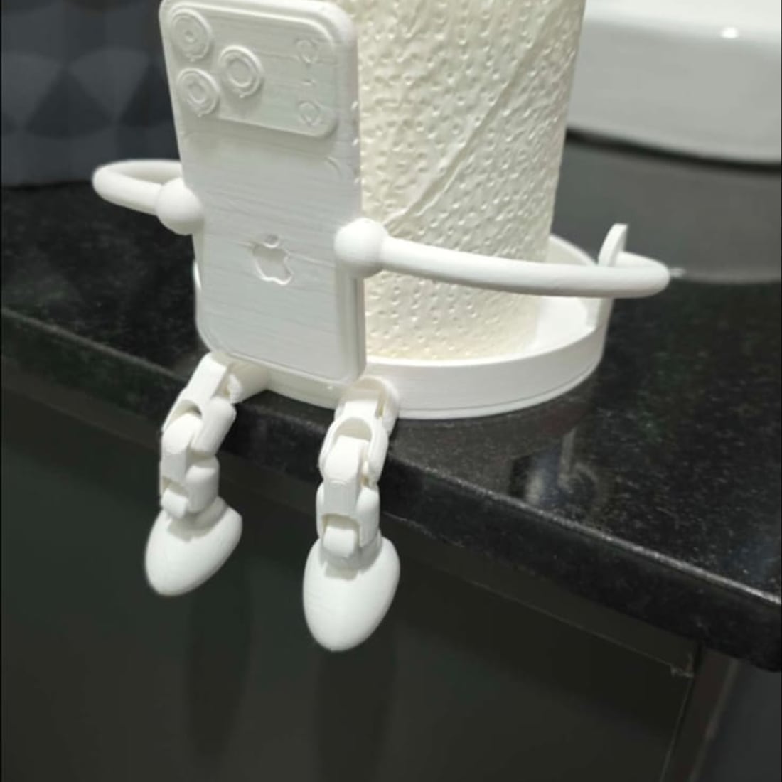 A white 3D-printed articulated robot figure sitting on a bathroom counter edge with its arms around a spare toilet roll, legs hanging over the side.
