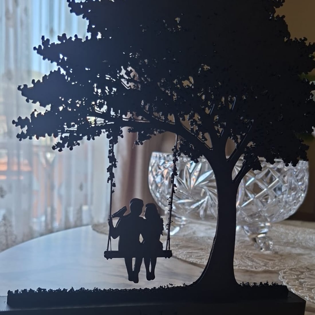 A black 3D-printed silhouette of a couple sitting on a swing beneath a full leafy tree, mounted on a Le Layers branded base, standing on a marble table.