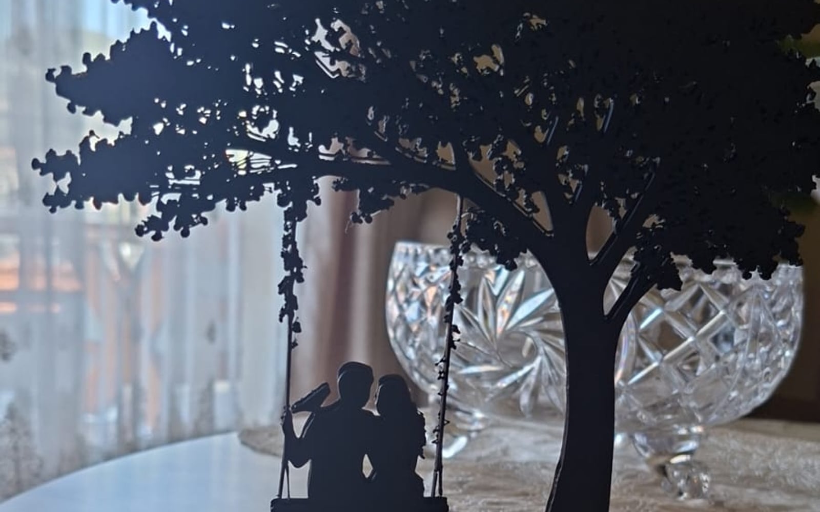A black 3D-printed silhouette of a couple sitting on a swing beneath a full leafy tree, mounted on a Le Layers branded base, standing on a marble table.